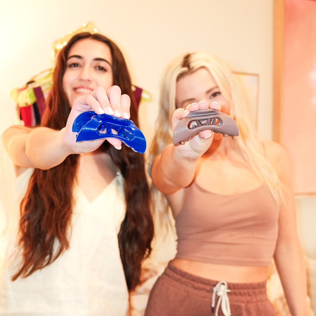 Two women holding Cozy Clip flexible claw clips—one Jelly cobalt blue and one Cove taupe—toward the camera in a bright indoor setting