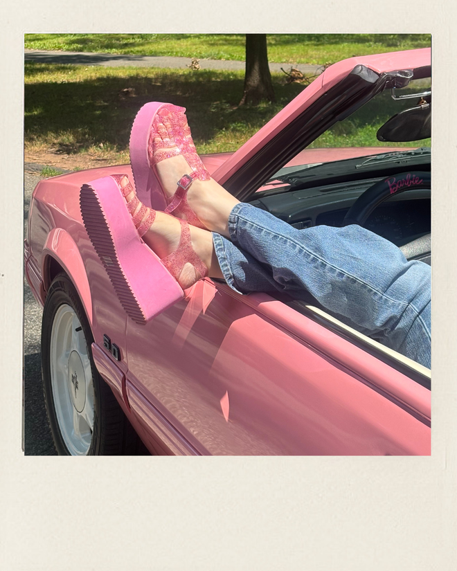 Person in blue jeans rests glittery pink jelly shoes on the door of a pink convertible