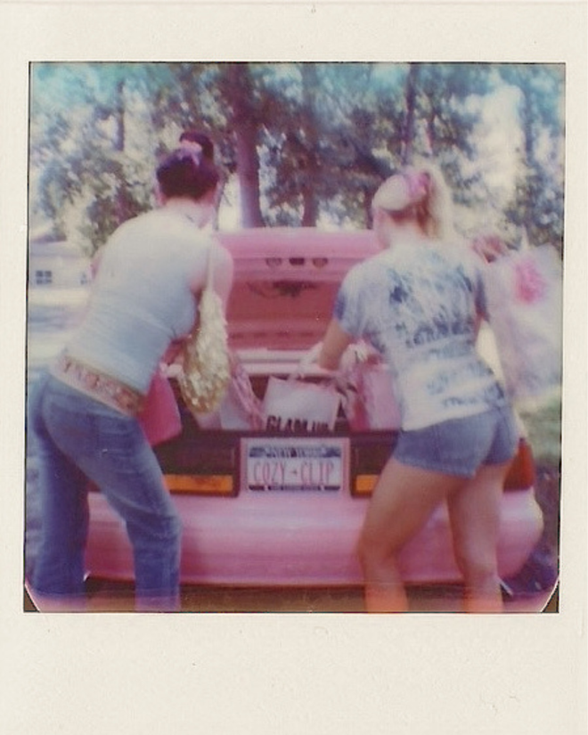 Two people stand beside a pink vehicle outdoors beneath trees in a vintage style photograph