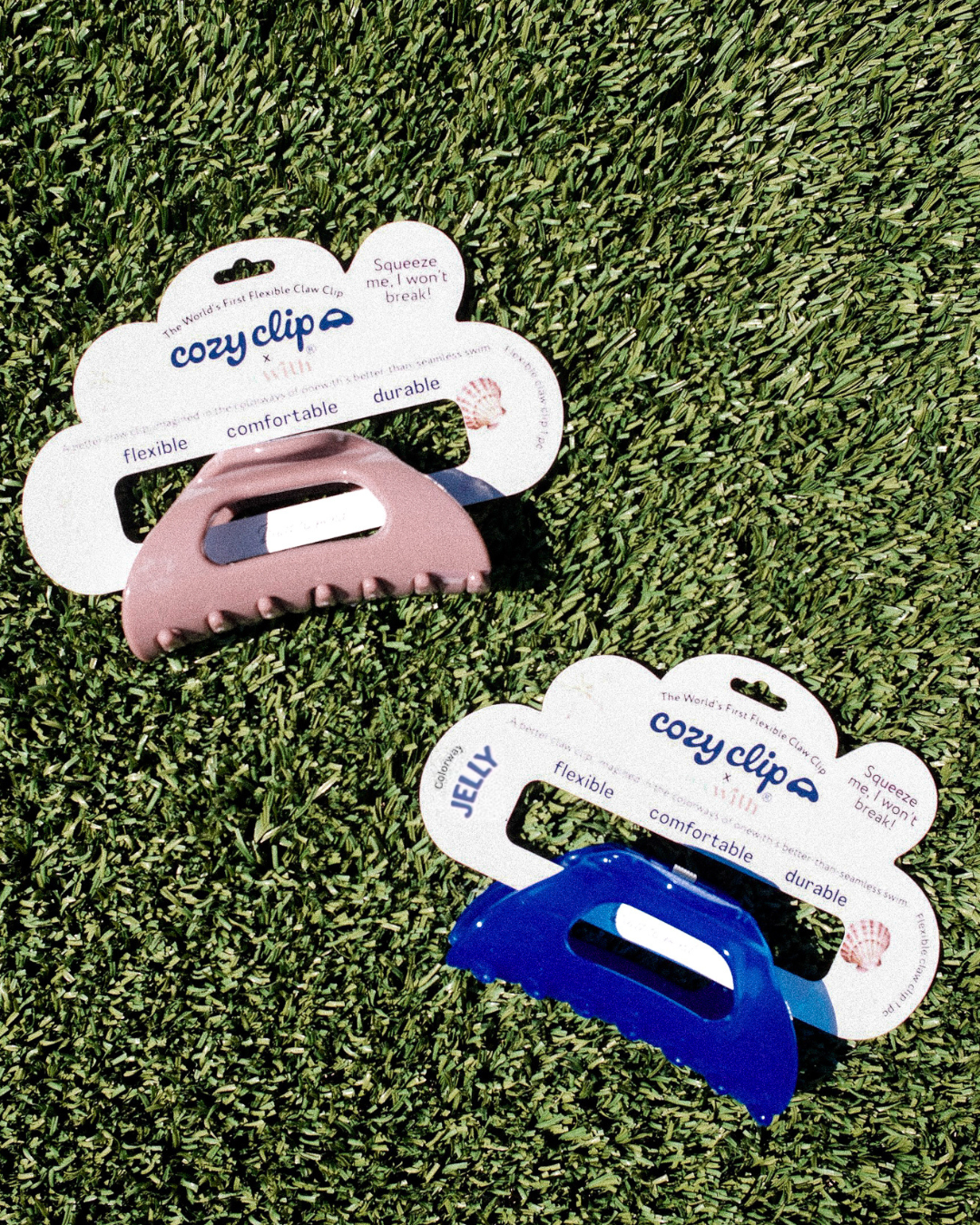 Top-down view of two Cozy Clip flexible claw clips in Jelly cobalt blue and Cove taupe on grass with packaging